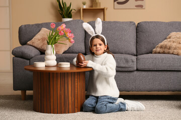 Cute little girl in bunny ears with sweet chocolate egg sitting on carpet in living room