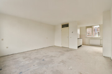 A spacious, empty room featuring white walls and a large window allowing natural light to flood in. The minimalistic design emphasizes simplicity.