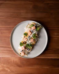 On a rustic wooden background, a plate of pasta with succulent salmon, crisp broccoli, and black olives. A comforting seafood meal in a cozy setting.