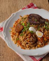 Traditional Uzbek plov pilaf with fragrant rice, tender beef, carrots, and spices, garnished with green onions, quail egg, and garlic. Served on a white plate over a rustic wooden table, top-down view