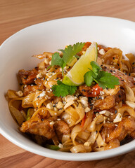Traditional Pad Thai with stir-fried rice noodles, tender chicken, vegetables, and peanuts, garnished with fresh cilantro and a lime wedge. Served in a white bowl on a wooden table, close-up view