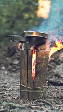 Close-up slow-motion view of a man's hand wearing a paracord bracelet reaching for a metal pot atop a burning Swedish torch to check the water temperature in a rugged outdoor survival atmosphere