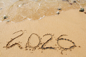 words 2026 and number written in sand on beach with waves on top. Happy New Year background