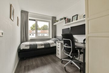 A cozy, modern bedroom featuring a comfortable bed, a study desk with monitors, and natural light streaming through large windows.