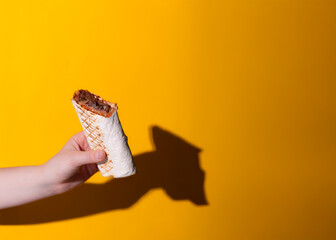 A horizontal photo of a hand holding a freshly wrapped shawarma with grilled meat, vegetables, and sauce, set against a bright orange background. Perfect for advertising or promotional use.