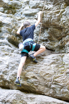 Young boy rock climbing with safety gear on a rugged outdoor cliff.