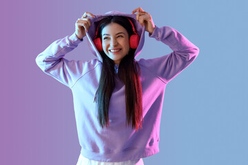 Stylish young woman in hoodie with headphones on color background