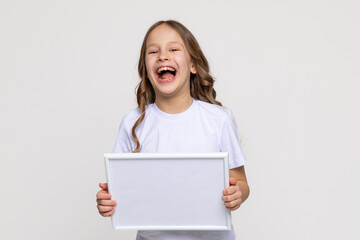 Fototapeta premium White empty banner. Laughing little school girl holds white blank paper. Young smiling woman show blank board. Close up female model portrait on white background. Mock up. Copy space.