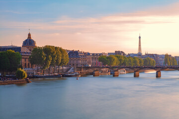 Fototapeta premium Cityscape of Paris and the Seine River at sunset, with the historic Institut de France on the left, the Pont des Arts bridge in the middle, and the iconic Eiffel Tower in the background.