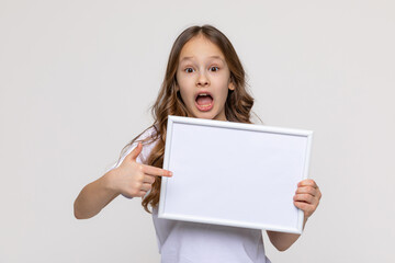 Fototapeta premium White banner. Surprised shocked little school girl in white t-shirt holds white blank paper, points index finger on empty board. Close up female model portrait on white background. Mock up. Copy space