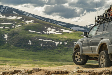 An off-road tourist car on mud tires stands against the background of a mountain that is covered with green grass and in places covered with white snow. A family car in the mountains. © ANDREY