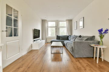 A bright and airy modern living room featuring a comfortable gray sofa, wooden flooring, and large windows that provide plenty of natural light.