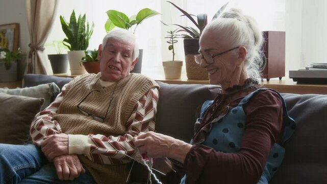 Elderly grey-haired woman sitting on couch, knitting with needles and chatting with male friend during daytime in nursing home