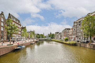 Fototapeta premium A charming view of a canal surrounded by traditional Dutch architecture and lush green trees, perfect for capturing the essence of Amsterdam's beauty.