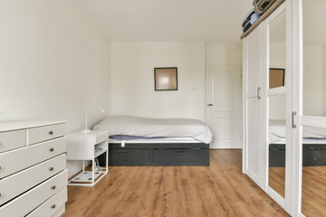 A stylish modern bedroom featuring a bed with soft linens, minimalist furniture, wood flooring, and a large mirror reflecting the serene atmosphere.