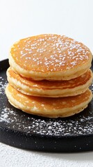 A stack of four golden brown pancakes dusted with powdered sugar sits on a dark gray plate against a bright white background.