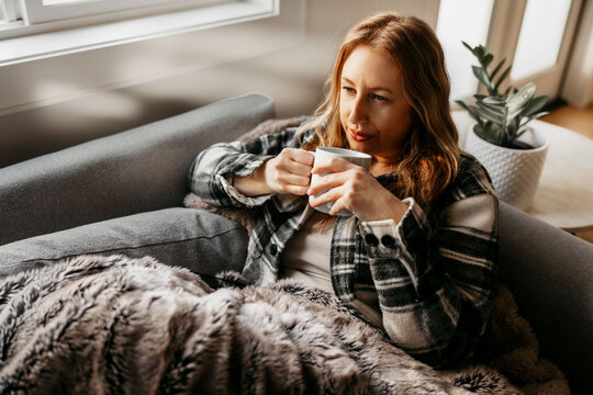 A woman in her 40s relaxes on the couch with a warm drink and blanket