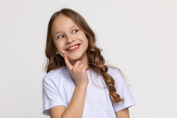 Joyful little girl in white T-shirt smiling widely and holding index finger to cheek, looking on...