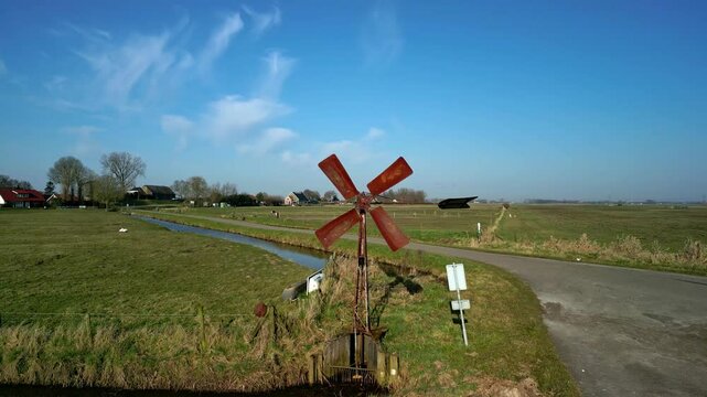 Drone view of an old windmill next to water canal and road in vast rural field under blue sky