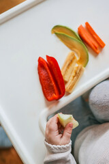 A baby's hand reaching for fresh vegetables and fruits on a white highchair tray. Includes sliced...