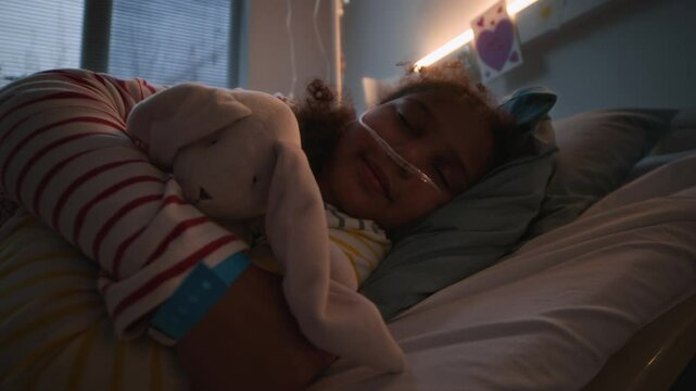 Cute little African American girl with nasal cannula sleeping on hospital bed hugging her favorite toy, resting while receiving medical treatment
