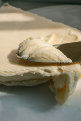 Close-up of a knife spreading fresh butter with soft curls forming on the blade. Warm natural light highlights the creamy texture, creating a rustic and appetizing food scene