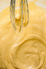 Close-up of a whisk mixing smooth yellow batter in a bowl. The thick, creamy texture of the mixture is highlighted by natural light, emphasizing the glossy and rich consistency of the dough