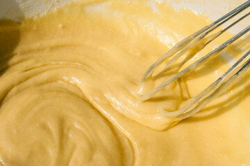 Close-up of a whisk mixing smooth yellow batter in a bowl. The thick, creamy texture of the mixture is highlighted by natural light, emphasizing the glossy and rich consistency of the dough