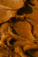 Close-up of creamy peanut butter spread with a rich, smooth texture. The golden-brown surface has visible swirls and ridges, showcasing its thick and glossy consistency