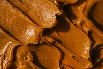 Close-up of creamy peanut butter spread with a rich, smooth texture. The golden-brown surface has visible swirls and ridges, showcasing its thick and glossy consistency