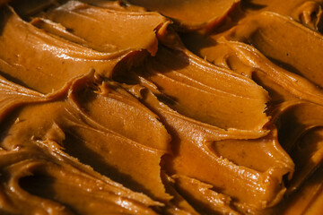 Close-up of creamy peanut butter spread with a rich, smooth texture. The golden-brown surface has visible swirls and ridges, showcasing its thick and glossy consistency