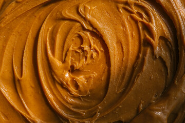 Close-up of creamy peanut butter spread with a rich, smooth texture. The golden-brown surface has visible swirls and ridges, showcasing its thick and glossy consistency