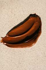A smear of glossy chocolate spread on a textured beige surface. The rich brown color contrasts with...