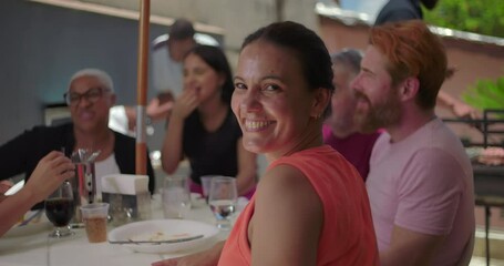 Woman smiling brightly at the camera during a lively family BBQ lunch, surrounded by friends and family enjoying an outdoor meal in a warm and cheerful atmosphere