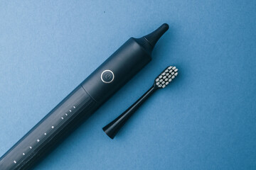 Black electric toothbrush with replaceable head on blue background. Modern rechargeable toothbrush for daily oral hygiene and dental care, minimalistic top view