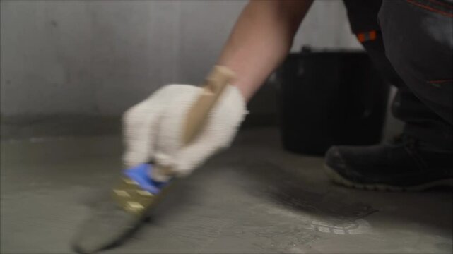 Professional construction worker applying liquid waterproofing membrane on concrete floor using specialized brush for basement renovation and protection