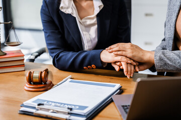 Two female lawyers in business suits hold hands at a wooden desk, offering comfort and encouragement. A wooden gavel and scales symbolize justice as they discuss corporate law, compliance