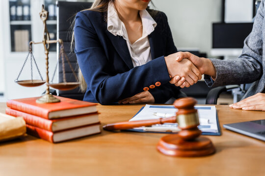 Two female lawyers in business suits shake hands at wooden desk, finalizing legal agreement. wooden gavel and scales represent justice as they discuss corporate law, compliance, dispute resolution.