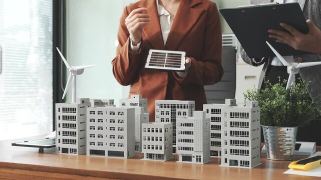 Two businesswomen sit at a desk in a meeting, discussing property development while building a model of city tower. real estate investments, zoning laws, sustainable energy solutions. net zero