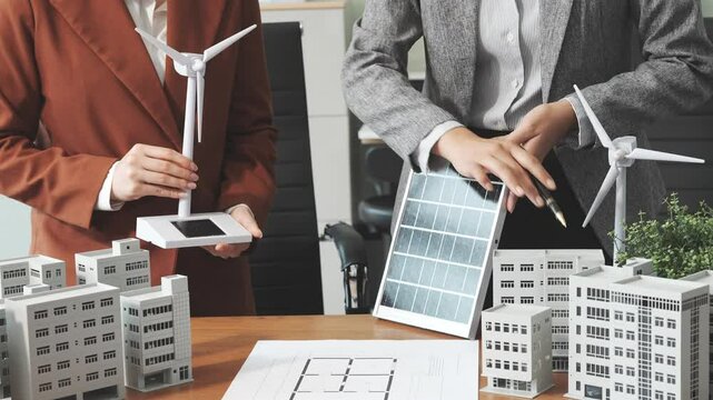 Two businesswomen sit at a desk in a meeting, discussing property development while building a model of city tower. real estate investments, zoning laws, sustainable energy solutions. net zero