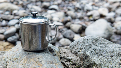 A stainless steel cup with its lid stands on a large rock amidst the tranquil flow of the river,...