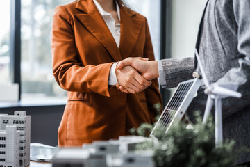 Two businesswomen shake hands at desk, finalizing deal on property development. real estate investments, zoning laws, sustainable energy solutions while working on city tower model, net-zero goals.