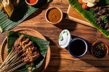 Traditional Indonesian satay served on banana leaves with peanut sauce, sambal, sweet soy sauce, and rice cakes on a rustic wooden table. A rich cultural and culinary food presentation