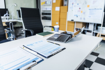 A modern office room with a white desk, laptop, financial chart, and inventory list. A whiteboard displays key data, alongside a pen, book, coffee cup, and black chair.