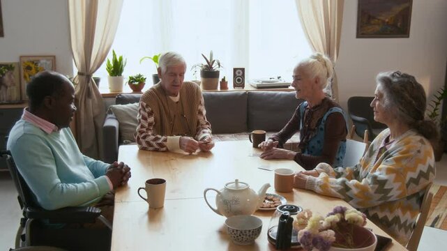 Elderly man giving cards to group of diverse friends sitting around wooden table with tea cups, playing game together during day in assisted living facility