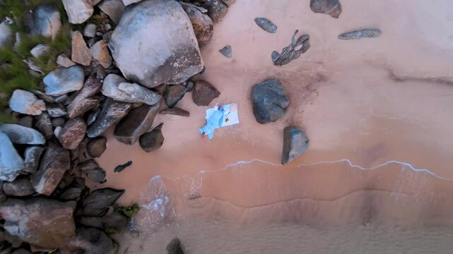 A serene scene of a woman enjoying a picnic on the sandy beach of El Gliaa in Bizerte, Tunisia. Surrounded by large rocks, it offers a peaceful moment by the sea.
