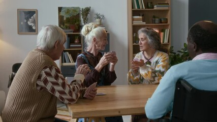 Group of multi-ethnic elderly friends chatting and laughing while playing card game in cozy living room of nursing home - Powered by Adobe