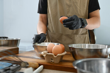 A man is cracking eggs into a bowl. He is wearing gloves and apron. There are several bowls on the table.