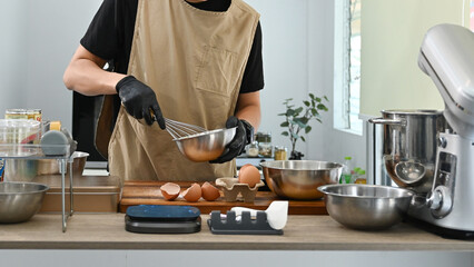 A man is making a cake in a kitchen. He is wearing apron and gloves. There are several bowls and spoons on the counter.