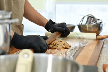 A man is making a dough on a wooden board. He is wearing black gloves. There are several bowls and cups on the table.